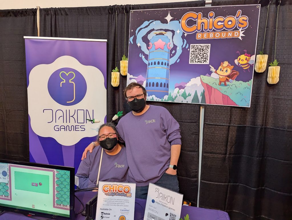 A photo of Nick and his wife at the Daikon Games booth at BostonFIG. They are wearing matching purple Daikon sweatshirts and black masks. She is wearing a headband with miniature daikon radishes on the ends of springs.
Behind them is a banner for Daikon Games and another for Chico's Rebound which shows the game's key art and a QR code.
In front of them is a table with a purple table cloth, upon which is a small TV running the game Chico's Rebound, and some various small signage.